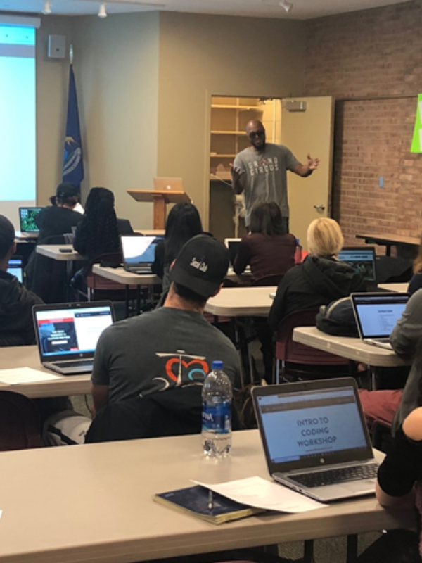 Rows of people with laptops open listen to an instructor about coding.